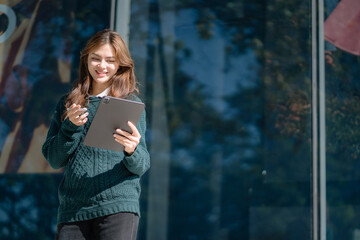 Smiling young woman leader entrepreneur, professional manager holding digital tablet computer using software applications standing on the street in big city on sky and office background.