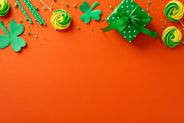 Saint Patrick's Day concept. Top view photo of green giftbox with ribbon bow meringue candies trefoils straws and sprinkles on isolated vibrant orange background with copyspace