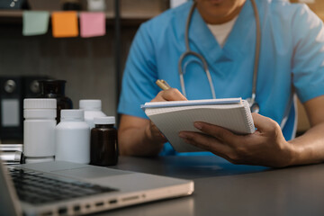 Male medicine doctor, physician or practitioner in lab room writing on blank notebook and work on laptop computer with medical stethoscope on the desk at hospital. Medic tech concept.