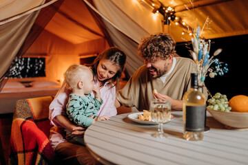 Happy family with lovely baby have dinner and spend time together in glamping on summer evening near cozy bonfire. Luxury camping tent for outdoor recreation and recreation. Lifestyle concept