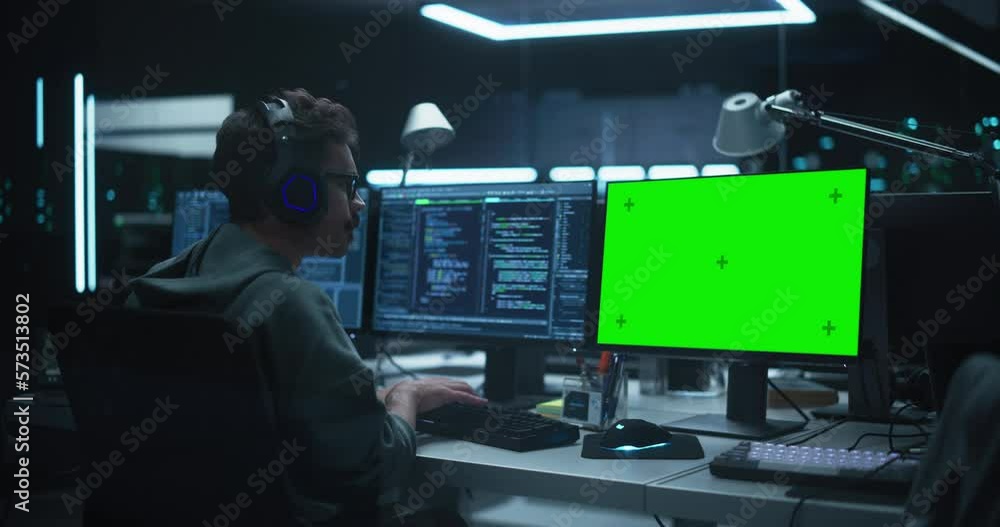 Aufkleber System Admin or Software Developer Working in a Dark Research Office Workplace on a Computer with Multiple Displays and Wearing Headphones. One of the Monitors is a Green Screen Mock Up Placeholder