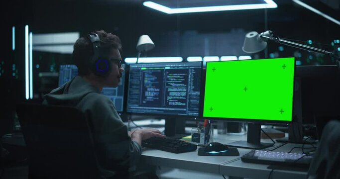 System Admin or Software Developer Working in a Dark Research Office Workplace on a Computer with Multiple Displays and Wearing Headphones. One of the Monitors is a Green Screen Mock Up Placeholder