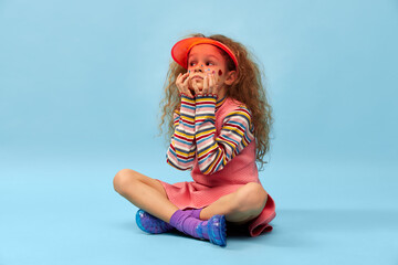 Calm playful kid. Little cute girl, child with curly hair posing in pink dress and cap over blue studio background. Concept of childhood, emotions, fun, fashion, lifestyle, facial expression