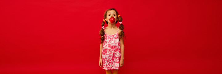 Little cute girl, child with curly hair posing, emotionally eating apple on bright red background. Concept of childhood, emotions, fun, fashion, lifestyle, facial expression. Banner. Copy space for ad
