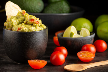 Close-up of black jar with guacamole on dark table with tomatoes, limes, spoon and avocados, black background, horizontal, selective focus, with copy space