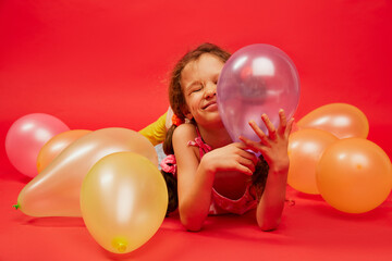 Little happy cute girl, child with curly hair posing with air balloons over bright red studio background. Concept of childhood, emotions, fun, fashion, lifestyle, facial expression