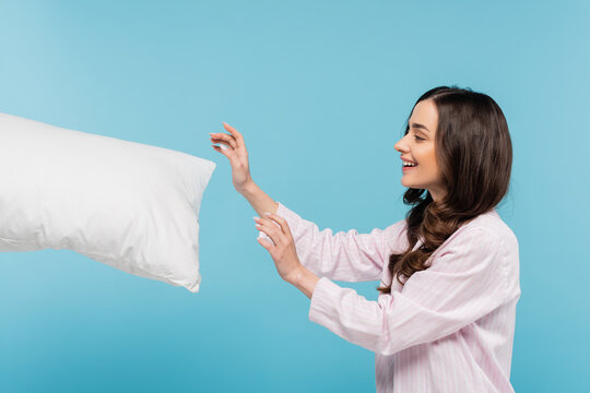 Side View Of Happy Young Woman In Sleepwear Reaching White Pillow In Air Isolated On Blue.