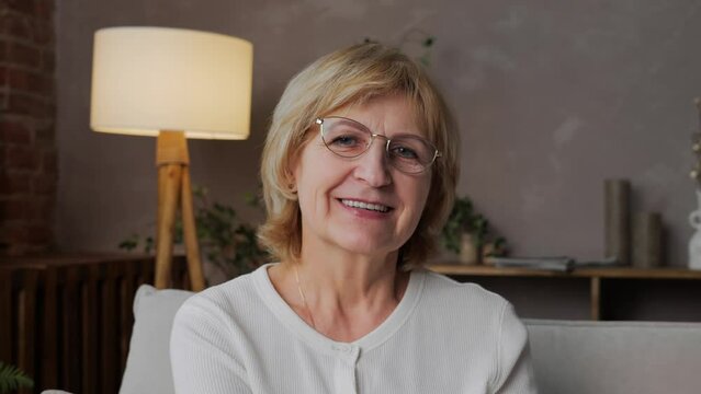Portrait of a happy 60-year-old blonde woman with glasses in a home interior looking into the camera. Kind and positive and beautiful adult woman freelance programmer. High quality 4k footage