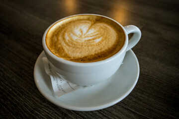 A white ceramic cup with hot coffee drink - cappuccino, americano with milk, latte - stands on a wooden table in a cafe bistro restaurant. Brown foam. Copy space. Selective focus. Breakfast drink.