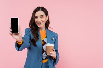 cheerful young woman in blue blazer holding smartphone with blank screen and paper cup isolated on pink.