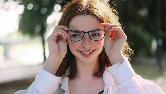 Portrait of pretty red haired young woman with bad poor vision wearing glasses and looking around enjoying clear focus image of the surrounding world outdoors Eye care eyesight and vision concept