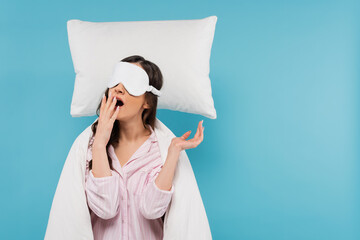 young woman in night mask yawning near levitating pillow isolated on blue.