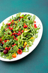 Summer delicious salad with arugula and fresh berries, red and black currants, sweet cherries and black mulberries, salad in an oval white plate on a green background
