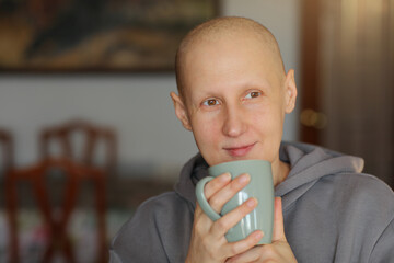 Hairless happy woman is holding cup of hot drink, coffee or tea. Bald female is having remission...