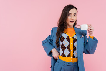 brunette woman in blue blazer holding cup of coffee isolated on pink.