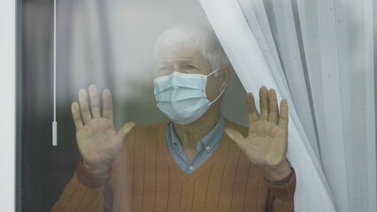 Portrait of old man wearing face mask looking out window, lonely and sad