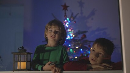 Two children looking out window, lamp and decorated Christmas tree