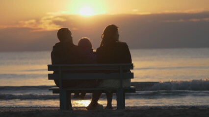 Portrait of family silhouettes sit on bench at sunrise, beach holiday