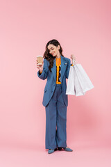 full length of pleased young woman in blue pantsuit holding shopping bags and coffee to go on pink.