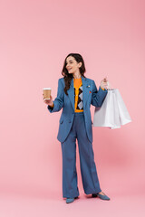 full length of happy woman in blue pantsuit holding shopping bags and coffee to go on pink.