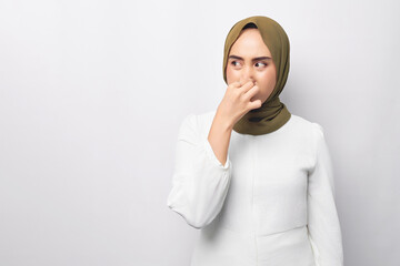 unhappy young beautiful Asian Muslim woman 20s wearing hijab feeling disgusted, holding her nose to avoid smelling foul, looking aside isolated on white background. People religious lifestyle concept - obrazy, fototapety, plakaty