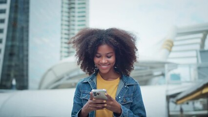 Happy young African American woman walking down the city street smiling using mobile phone chatting online with friends, Female in jean jacket walks down the street using social media on smartphone - Powered by Adobe