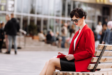 Business, paperwork and people concept - confident business woman reading and writing notes in notepad sitting on city bench. Thoughtful female wearing in red blazer and leather skirt looking forward