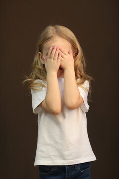 little blond baby girl in white t-shirt with copy space close her eyes with hands close up photo on dark brown wall background