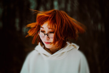 Portrait of a beautiful ginger Asian woman with glasses and a white hoodie, who waves her hair in a funny way against the background of a dark park. Lifestyle.