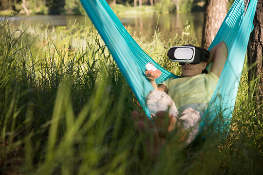 middle aged woman playing game in virtual reality glasses in a hammock relaxed outdoors. copy space.