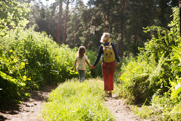 Naklejka na ściany i meble mom and and child daughter with backpack back to the viewer walking in the forest. Staycations, hyper-local travel, family outing, getaway, natural environ. Concept of friendly family. 