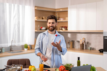 Obraz premium Glad attractive adult caucasian man in casual cooks dinner, juggles tomatoes in modern kitchen interior
