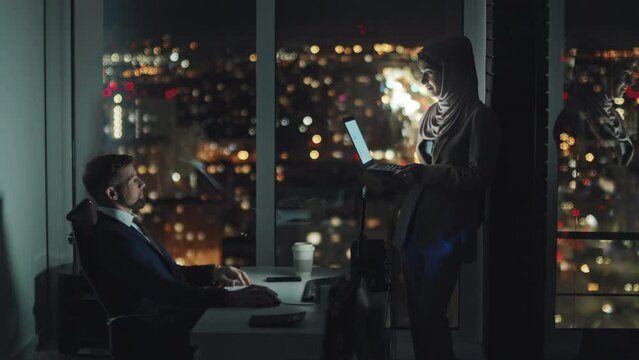 Full shot of Muslim female office worker in Islamic headscarf, holding laptop standing in dark office and asking Caucasian male colleague sitting at desk for advice on presentation, late at night