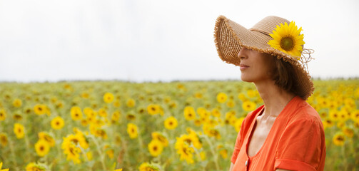 middle aged woman in an orange dress and a straw hat with a sunflower in a field with sunflowers.  ...