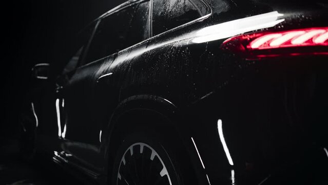 Aesthetic Cinematic Footage with a Modern Black Electric Luxury Vehicle. Close Up of an SUV with Glossy Wet Surface Standing in a Dark Detailing Studio Interior, Getting Dry After a Thorough Car Wash