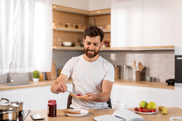Fototapeta na wymiar Breakfast and good morning. Happy adult caucasian man in white t-shirt makes sandwich with jam at table