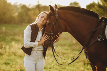 Young beautiful female jockey strokes and hugs the horse's head and prepares for the competition. Jumping training in the meadow in summer evening.