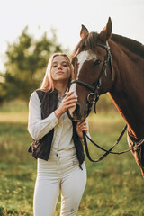 Obraz premium Young beautiful female jockey strokes and hugs the horse's head and prepares for the competition. Jumping training in the meadow in summer evening.