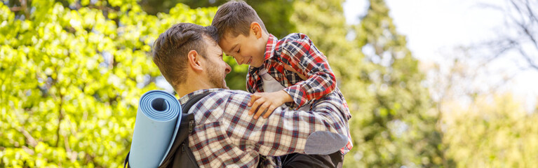 Cheerful Caucasian father with backpack holding small son on hands in park on summer day.