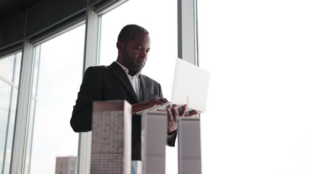 Smiling african american business man real estate agents in black stylish formal suit standing with laptopnear 3d model skyscrapers of city architecture design at office with panoramic city view.