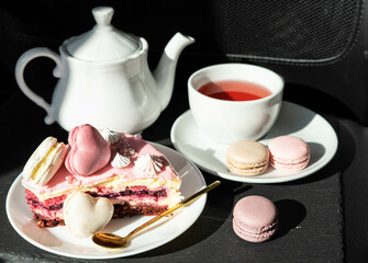 Pink cake with heart shaped macaroons , red tea, white kettle and gold spoon on a black background. Natural light, sunny day