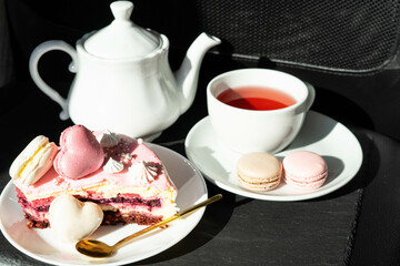 Pink cake with heart shaped macaroons , red tea, white kettle and gold spoon on a black background. Natural light, sunny day
