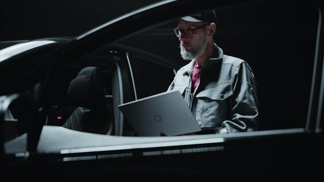 Portrait of a Dealership Car Mechanic Ananlyzing Diagnostics Results on a Modern Electric Vehicle. Software Developer Working on Laptop Computer, Investigating Settings on a Modern Sustainable EV
