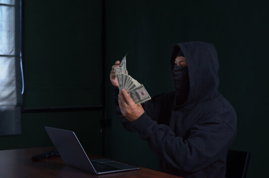 hacker in black hoodie and face with cover mask holding banknote in dark room, there are laptop and gun on table,concept of cyber crime