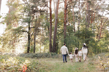 Naklejka na ściany i meble Back view of mother, father with three kids are walking, having fun in autumn forest. Family holding children hands enjoying time together at nature background. Happy parenthood and childhood concept