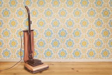 Retro styled image of a vintage brown vacuum cleaner in a room with floral wallpaper and wooden...