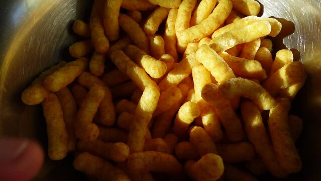 White hand grabs a large load of peanut flips puffs . Chips snack in a silver bowl ready to eat . Unhealthy but beautiful yummy food .