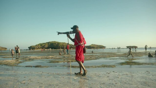 Masai Warrior In Red Traditional Clothes Standing At The Beach Holding A Wooden Pole And Pointing Direction In Watamu, Kenya. wide