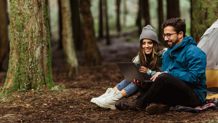 Cheerful millennial caucasian couple drink hot coffee in cold forest, enjoy adventure, sit in tent, typing on laptop