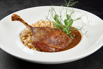 Classic dish french cuisine - roasted duck leg in porto sauce with garnish on black background. Cooked duck leg with pearl barley on white plate on dark concrete table. Duck confit in minimal style.